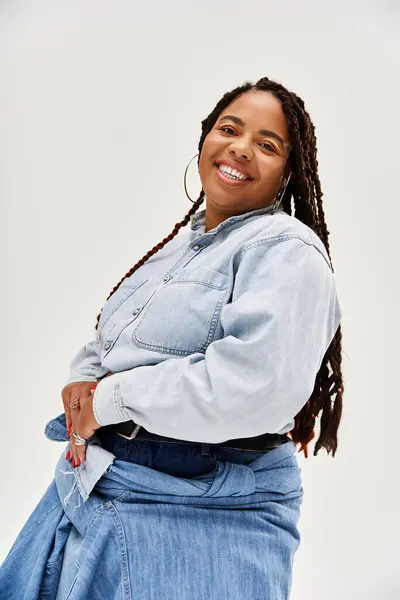 Una donna sicura di sé posa con un sorriso, mostrando le sue trecce ed elegante vestito di denim in uno studio. — Foto stock