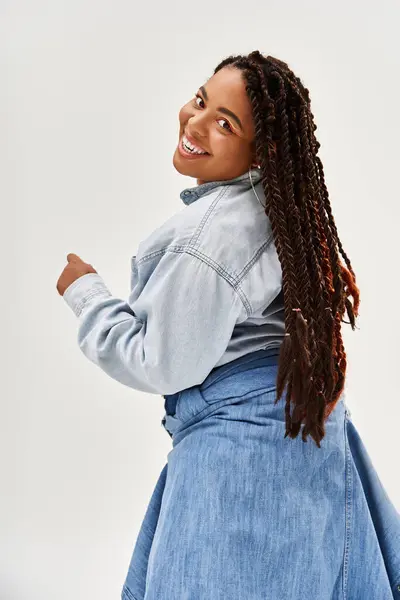 Uma jovem radiante em tranças posa confiante em sua roupa jeans contra um pano de fundo cinza. — Fotografia de Stock