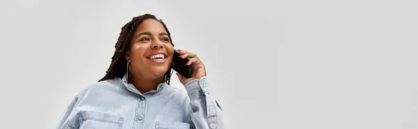Una mujer afroamericana bonita usando un traje de mezclilla sonríe brillantemente mientras está en una llamada telefónica, pancarta - foto de stock