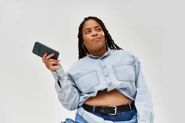 Una donna fiduciosa posa in una camicia di jeans ritagliato e jeans su uno sfondo grigio, trasudando gioia. — Foto stock