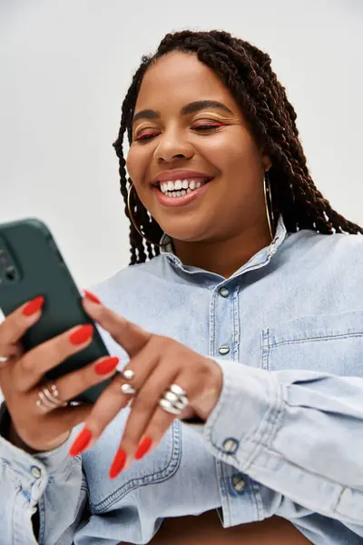 A cheerful African American woman in a denim outfit enjoys looking at her phone, smiling brightly. — Stock Photo