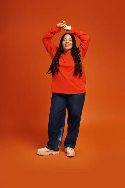 Une belle femme noire avec des tresses pose ludique dans une tenue colorée sur fond orange. — Photo de stock
