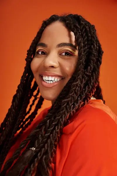 Une belle femme afro-américaine avec des tresses élégantes sourit sur un fond orange vif. — Photo de stock