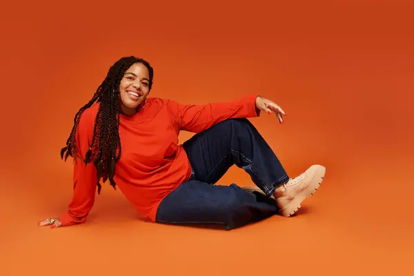 A joyful African American woman with braids strikes a stylish pose on a vibrant orange background. — Stock Photo