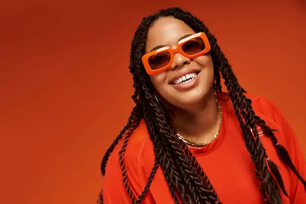 Una alegre mujer negra con trenzas muestra su atuendo de moda y sus gafas de sol sobre un fondo naranja. - foto de stock