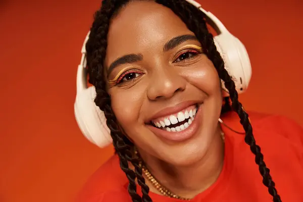 A joyful African American woman in a bright headphones poses against a vibrant orange background. — Stock Photo