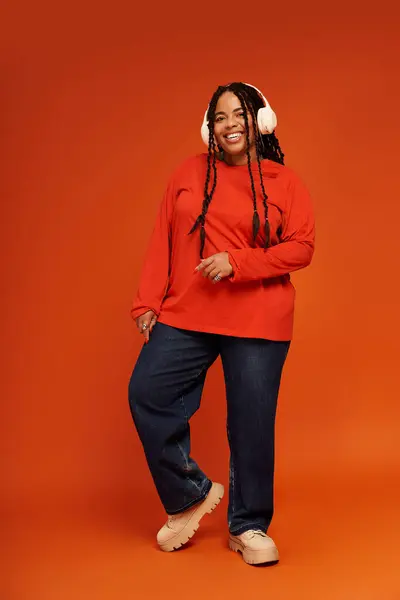 A stylish Black woman with braids smiles in a casual outfit with headphones against an orange background. — Stock Photo