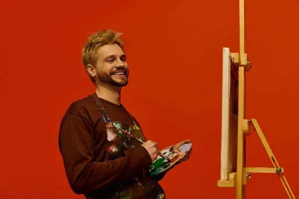 A handsome young man stands joyfully as he paints on an easel, expressing his creativity. — Stock Photo
