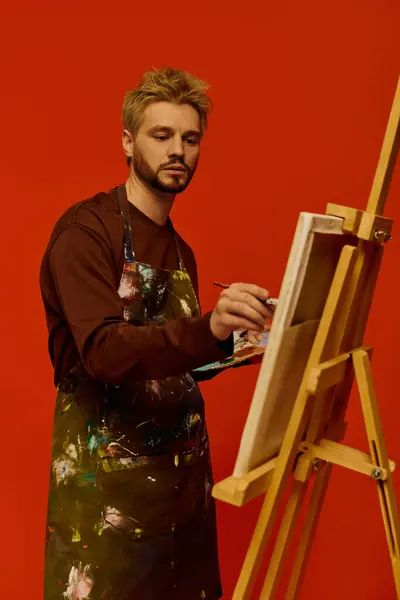 Joven guapo inmerso en la pintura en un caballete, rodeado de un fondo rojo vivo. - foto de stock