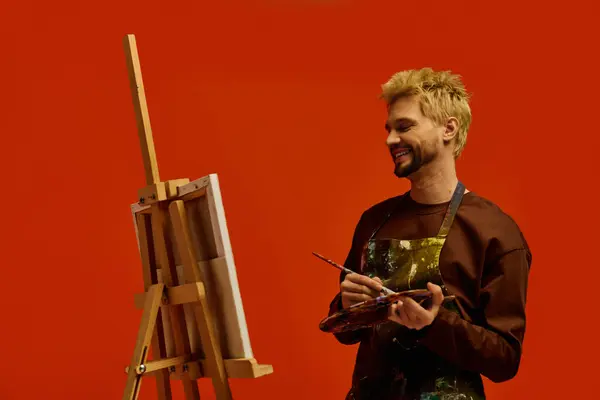 Handsome young man smiles while holding paintbrush and palette in artistic studio. — Stock Photo