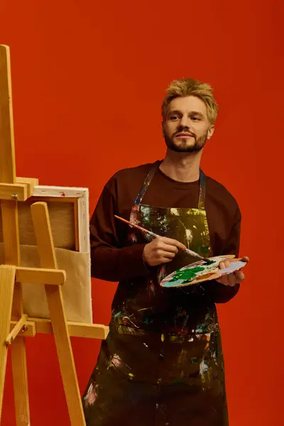 Handsome young artist engages in painting, mixing colors on a palette in a lively studio. — Stock Photo