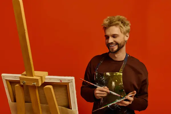 Handsome young man smiles while painting on an easel in a vibrant studio filled with creativity. — Stock Photo
