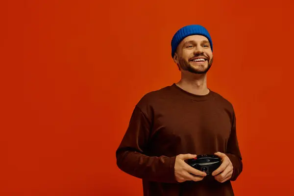 Bello giovane uomo tiene controller di gioco, sorridendo ampiamente su uno sfondo arancione vibrante. — Foto stock