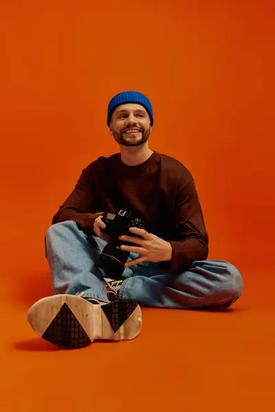 Giovane uomo siede gambe incrociate, sorridente con una macchina fotografica in un caldo studio colorato. — Foto stock