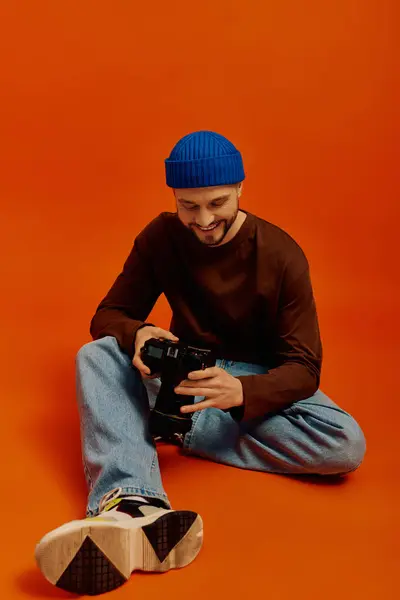 Un joven con un gorro azul sonríe, examinando su cámara con un fondo naranja brillante. - foto de stock