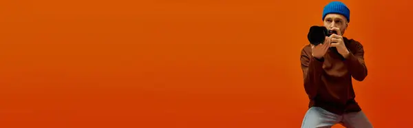 Handsome young man poses confidently with a camera against a bold orange backdrop. — Stock Photo