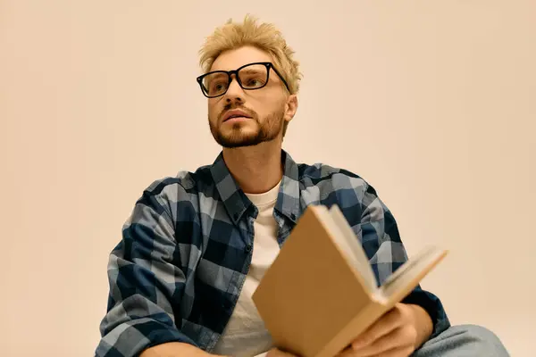 Captivating young man with blonde hair and glasses enjoys reading a book in a serene indoor setting. — Stock Photo