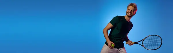 Un bel giovane uomo sorride ampiamente tenendo in mano una racchetta da tennis e una palla, pronto a giocare. — Foto stock