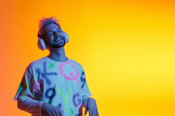 Bello giovane uomo che indossa cuffie sorride brillantemente mentre si gode la musica in un ambiente colorato. — Foto stock