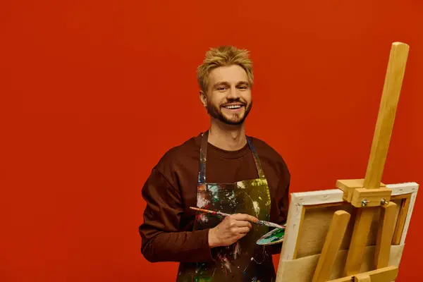 Handsome young man showcases his artistic talent by painting on an easel, smiling joyfully. — Stock Photo