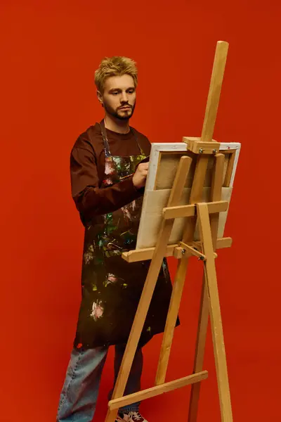 Joven guapo se para en el caballete, pintando apasionadamente sobre un atrevido telón de fondo rojo. - foto de stock