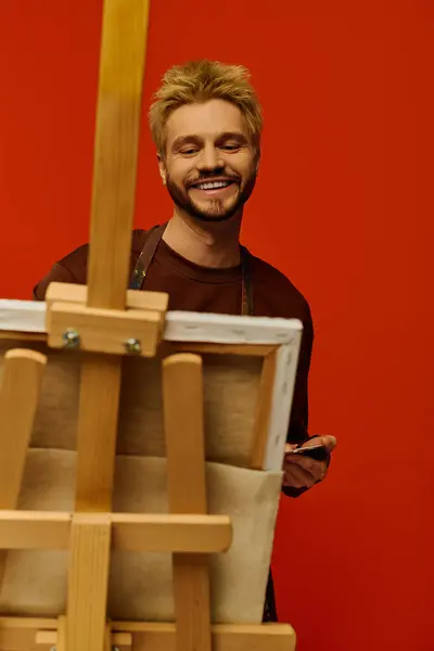 Un joven alegre se para junto a un caballete, mostrando su espíritu artístico en un colorido estudio. - foto de stock