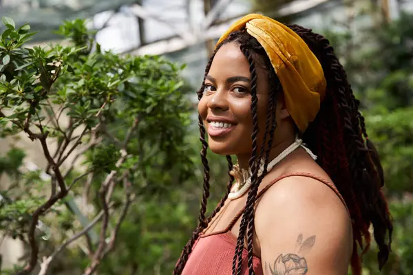 A cheerful African American woman with braids smiles amid greenery in a cozy greenhouse. — Stock Photo