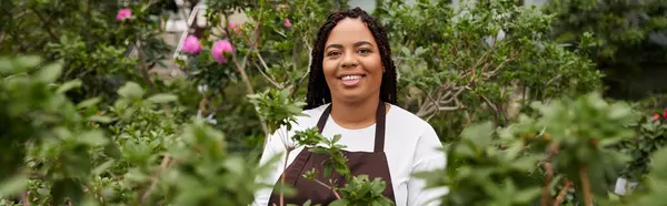 Una mujer afroamericana con trenzas nutre alegremente diversas plantas en un invernadero soleado, pancarta - foto de stock