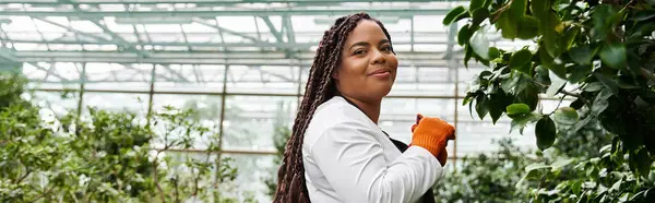 An African American woman with braids smiles while tending to plants in a vibrant greenhouse, banner — Stock Photo