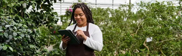 An african american woman with braids enjoys caring for plants in a bright greenhouse, banner — Stock Photo