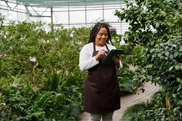 An African American woman with braids takes notes in a vibrant greenhouse. — Stock Photo