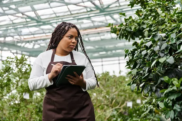 An african american woman with braids takes detailed notes on plants in a vibrant greenhouse. — Stock Photo