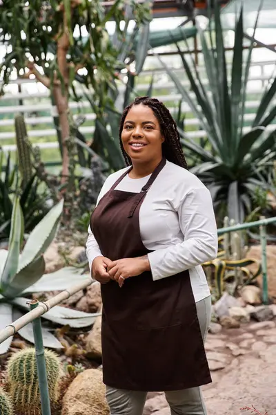 An African American woman with braids tends to her plants in a lush greenhouse, radiating joy. — Stock Photo