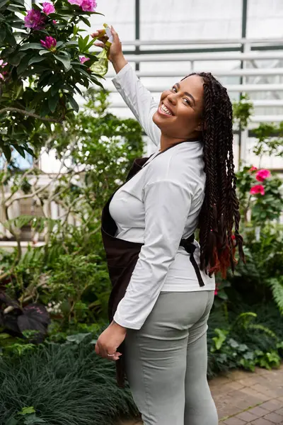 An African American woman with braids smiles while tending to blossoming plants in a greenhouse. — Stock Photo