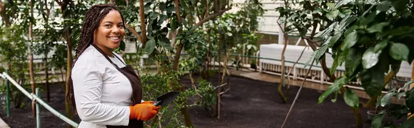 Uma mulher afro-americana com tranças tende a plantar em uma estufa exuberante, cultivando sua paixão, bandeira — Fotografia de Stock