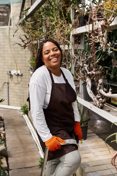 A smiling African American woman with braids stands in a greenhouse, tending to lush plants. — Stock Photo