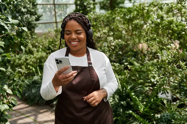 Una mujer afroamericana con trenzas sonríe en su teléfono en un exuberante invernadero. - foto de stock
