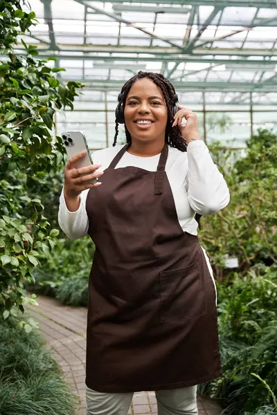 An African American woman with braids smiles while tending plants in a vibrant greenhouse habitat. — Stock Photo