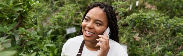A smiling African American woman with braids chats on her phone in a plant-filled greenhouse. — Stock Photo