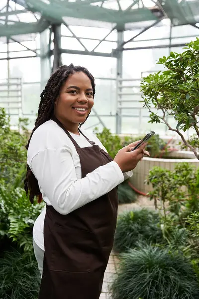 An African American woman with braids smiles as she tends to her plants in a greenhouse. — Stock Photo
