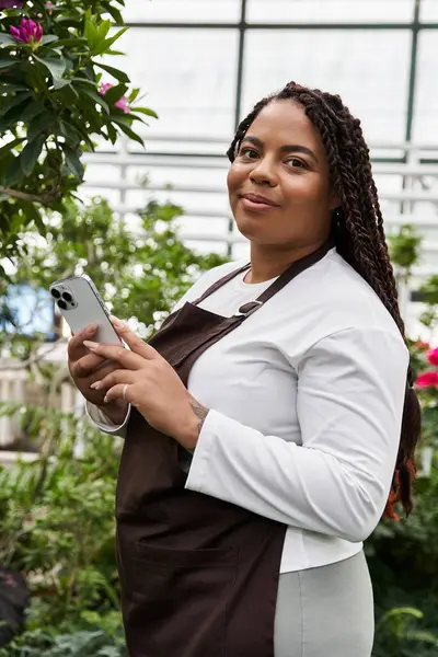 An African American woman with braids stands in a vibrant greenhouse, tending to plants. — Stock Photo