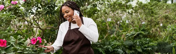 A happy Black woman chats on her phone while caring for vibrant flowers in a greenhouse, banner — Stock Photo