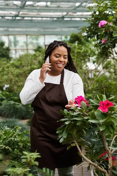 An african american woman with braids nurtures plants while talking on her phone in a greenhouse. — Stock Photo