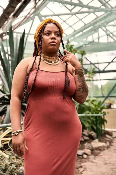 Una mujer confiada con trenzas posa graciosamente en un invernadero entre varias plantas. - foto de stock