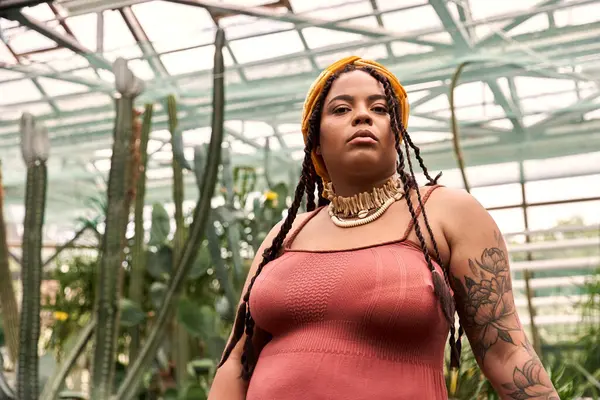 In a vibrant greenhouse, a beautiful woman with braids stands confidently among the greenery. — Stock Photo