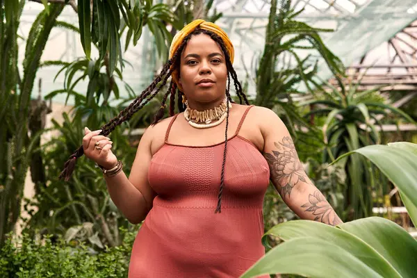 Una bella donna afroamericana con trecce posa tra piante lussureggianti in una serra luminosa. — Foto stock