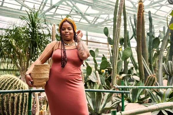 A pretty African American woman in a dress poses among lush greenery in a greenhouse. — Stock Photo
