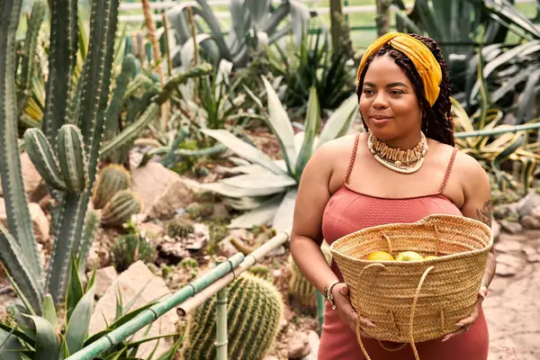 A pretty African American woman stands in a greenhouse, holding a basket of fruits with joy. — Stock Photo