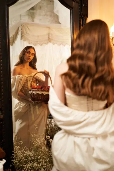 A young woman in an exquisite vintage dress holds a basket while gazing into a lavish mirror. — Stock Photo