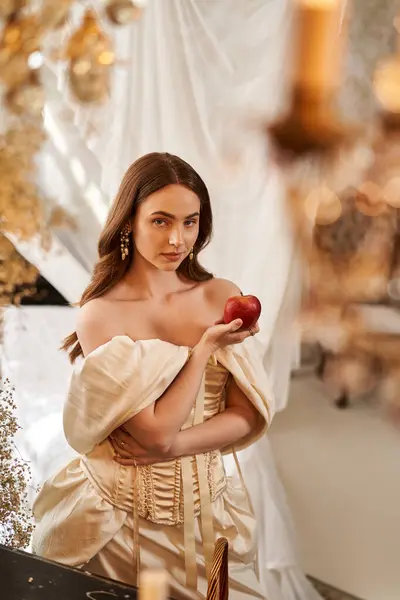 A lovely young woman in a vintage gown holds a red apple against a decorative backdrop. — Stock Photo